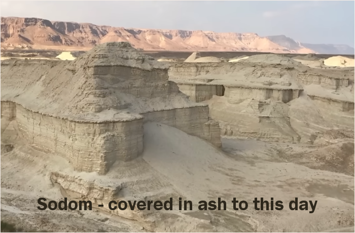 A panoramic view of the geological formations of Sodom, characterized by ash-covered cliffs and a barren landscape, with mountains in the background.