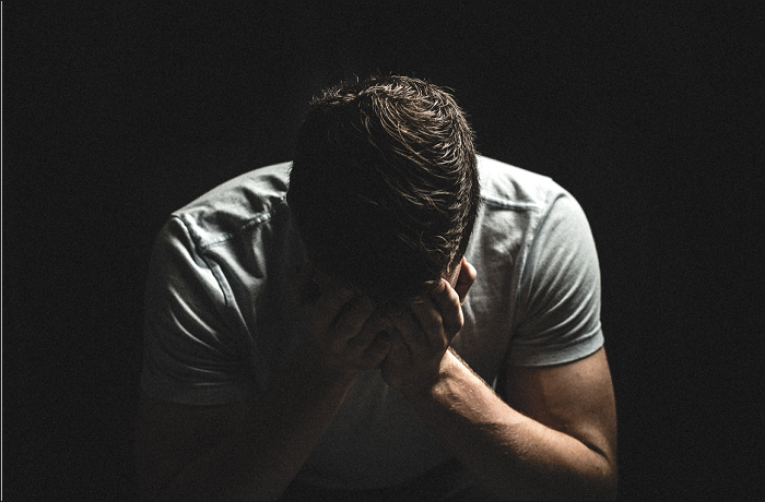 A person with their head down and hands covering their face, expressing sorrow or distress, in a dimly lit setting.