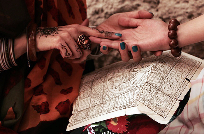 Two hands connecting, one adorned with henna and accessories, the other with colorful nail polish, while resting on an ornately designed piece of paper.