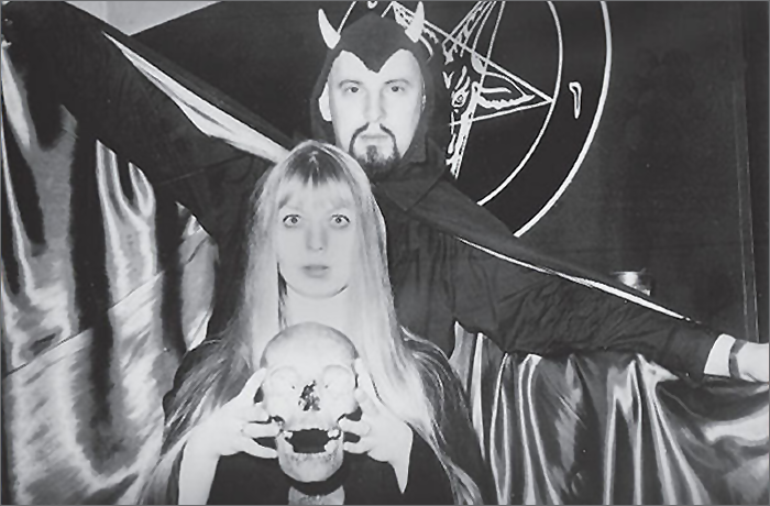 A man dressed in a devil costume standing behind a woman holding a skull, set against a dark background with occult symbols.
