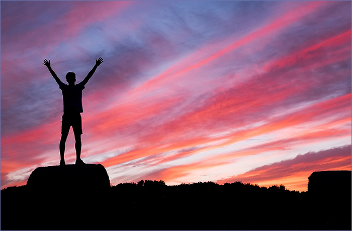 Silhouette of a person with arms raised, standing on a haystack against a colorful sunset sky with pink and orange hues.