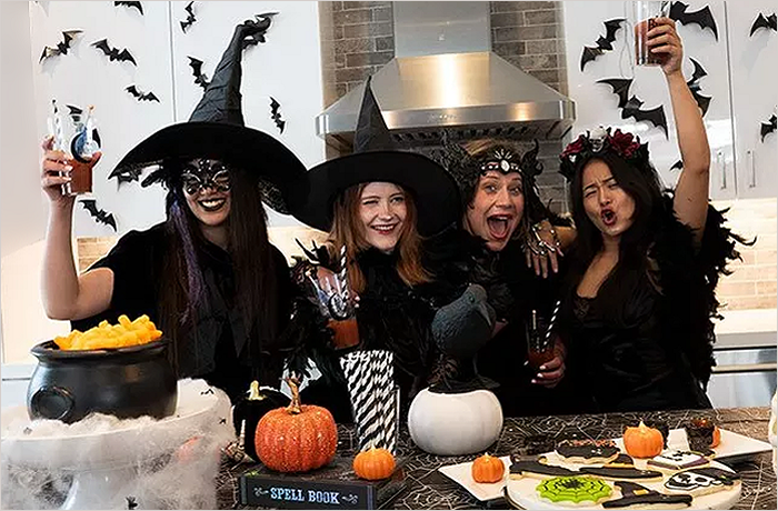 Four women dressed in Halloween costumes celebrating at a decorated kitchen, featuring a cauldron, pumpkins, and Halloween-themed treats.